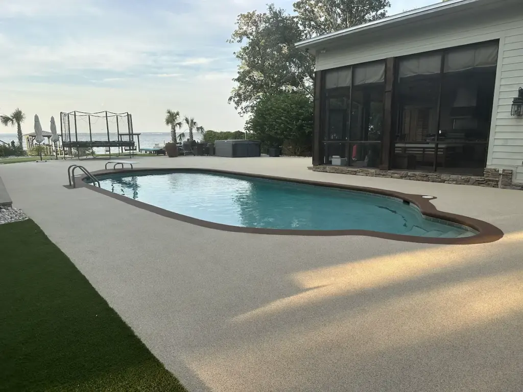 "A backyard swimming pool featuring a seamless light tan deck with a dark brown border, located next to a screened-in porch and overlooking a body of water.