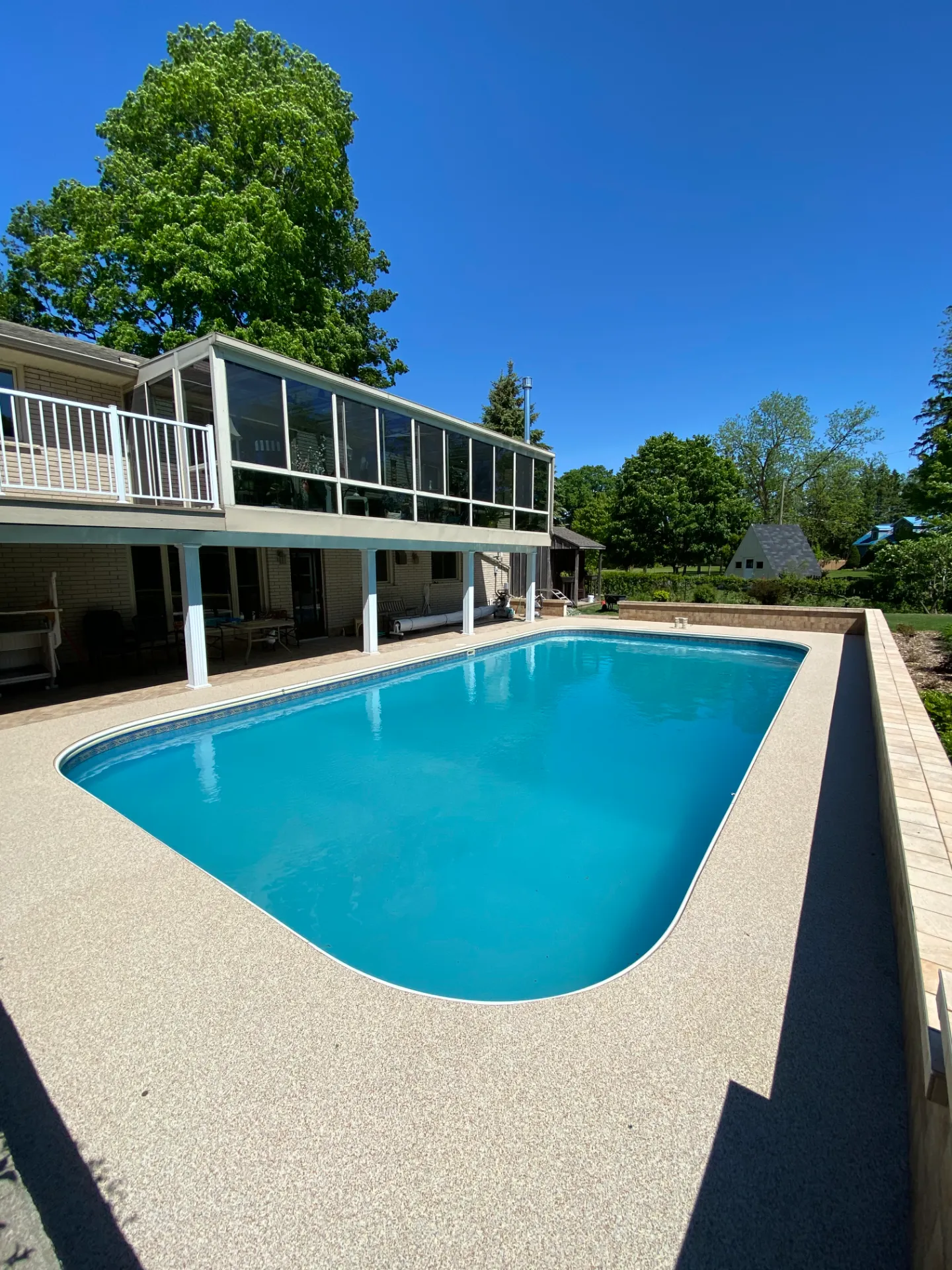 After photo of a backyard swimming pool featuring a newly resurfaced, seamless light tan aggregate pool deck. The bright turquoise pool sits next to a house with a raised sunroom and white pillars under a clear blue sky.