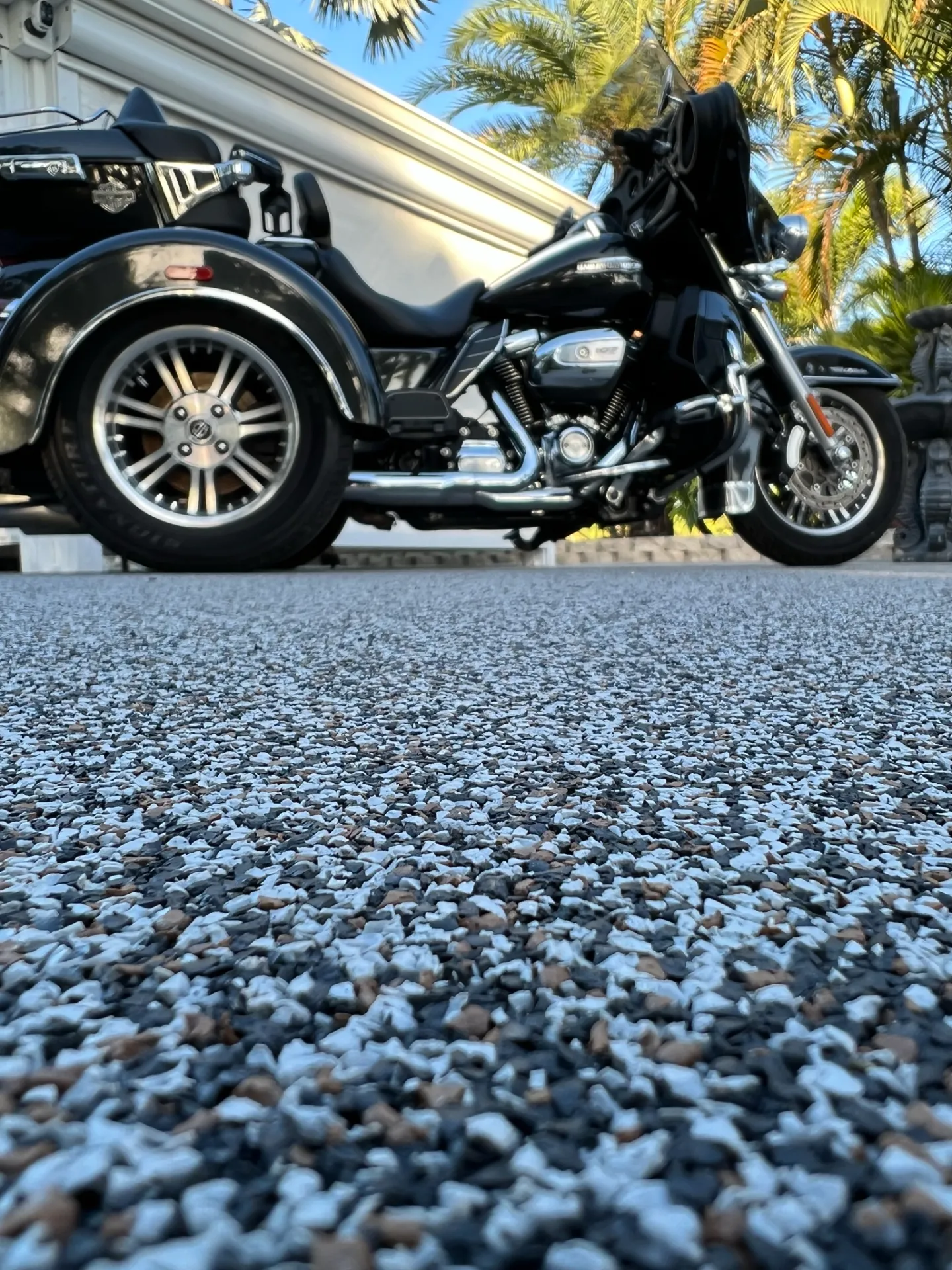 Close-up, low-angle view of a grey, black, and brown speckled resin rock surface, with a black three-wheeled motorcycle parked in the background in front of a white building and palm trees.