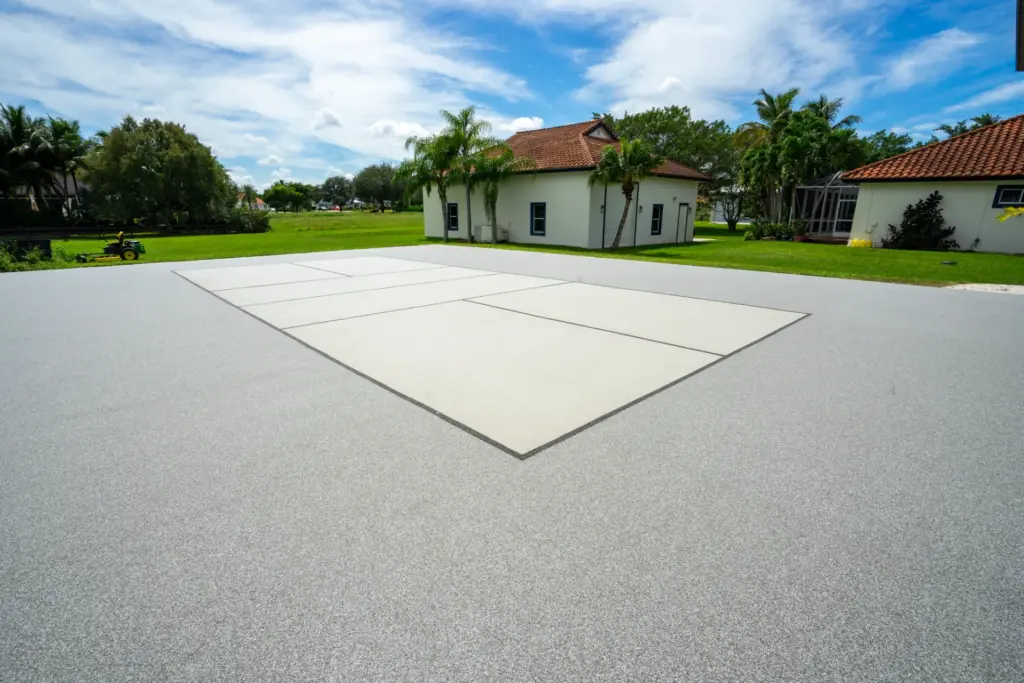 A large outdoor sports court featuring a seamless, grey speckled surface. The center playing area is a lighter shade marked with dark boundary lines, surrounded by a green lawn, palm trees, and white buildings with terracotta tile roofs under a bright blue sky.