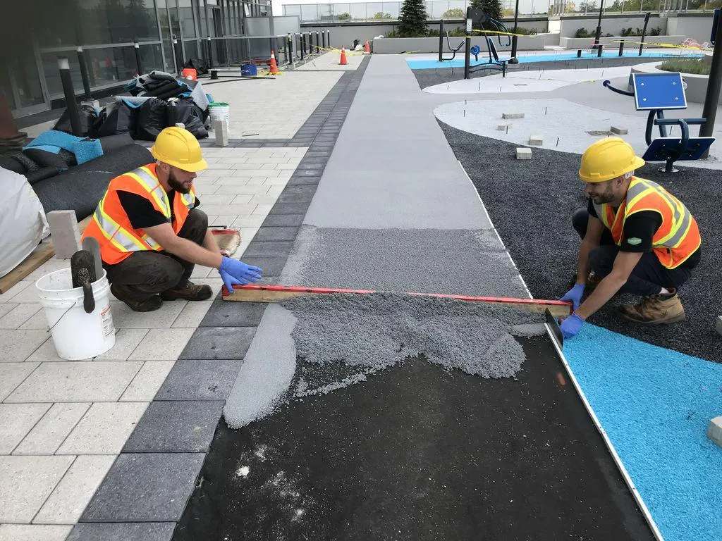 Two construction workers wearing yellow hard hats and safety vests kneel on the ground, using a long straight edge to smooth and level a fresh layer of grey rubber surfacing. The outdoor construction site features a paved stone walkway and adjacent sections of bright blue and black surfacing.