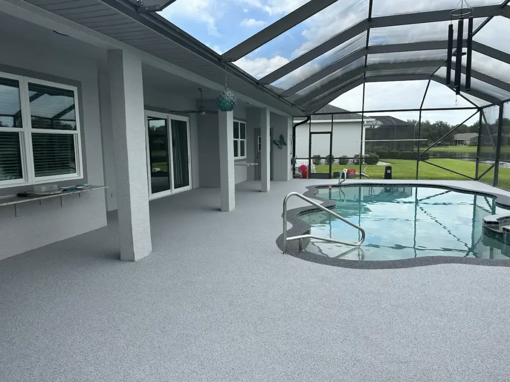 A screened-in outdoor patio and swimming pool area featuring a seamless, light grey speckled surface coating across the entire deck. The freeform pool is highlighted by a darker grey textured border, attached to a light grey house overlooking a backyard pond.