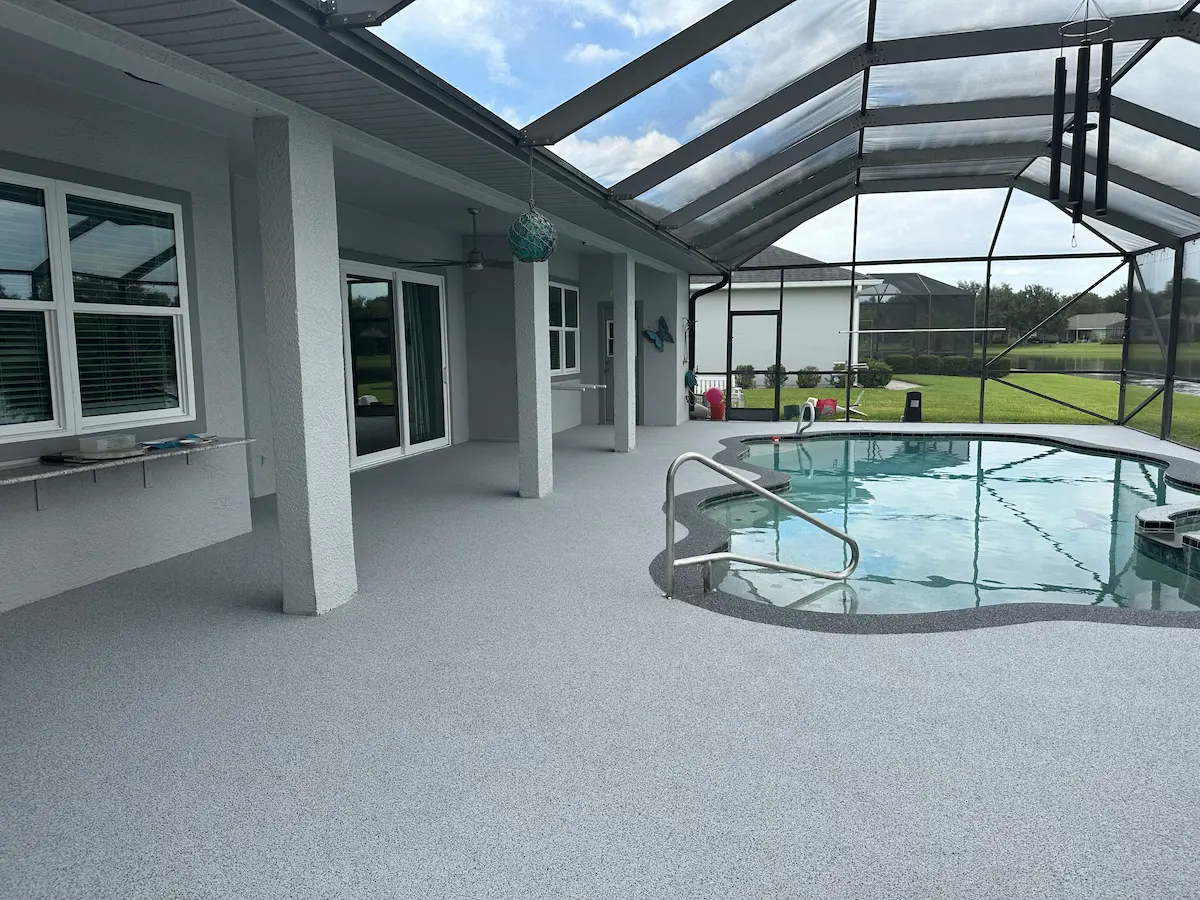 A screened-in outdoor patio and swimming pool area featuring a seamless, light grey speckled surface coating across the entire deck. The freeform pool is highlighted by a darker grey textured border, attached to a light grey house overlooking a backyard pond.