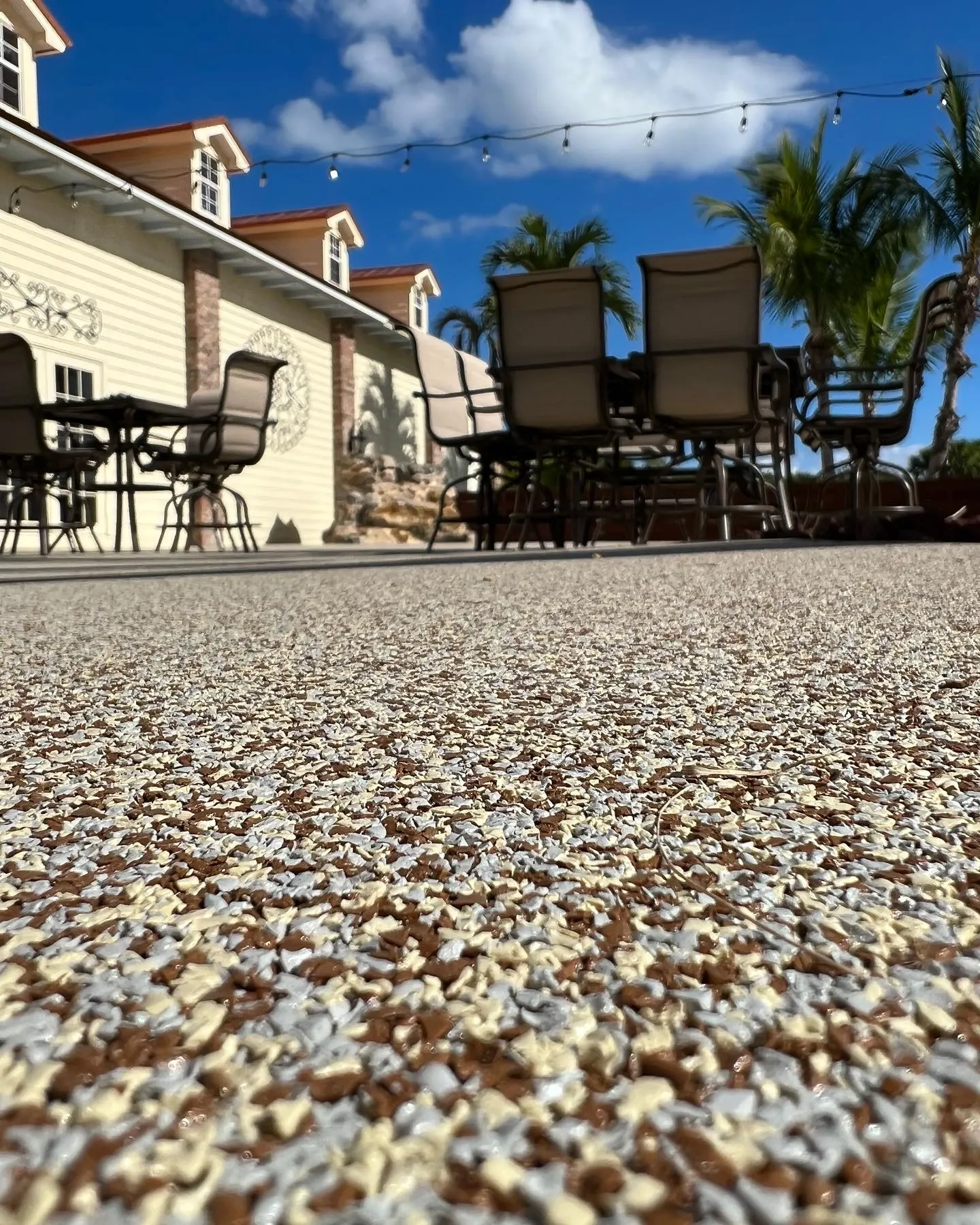 Close-up low-angle view of a speckled white and brown rubber stone patio surface, with outdoor dining furniture, a light yellow building, and palm trees in the background under a clear blue sky.